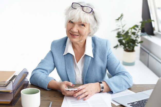 Femme d'âge moyen confiante moderne dans la soixantaine ayant une petite pause, assise sur son lieu de travail pour vérifier les nouvelles ou faire défiler les médias sociaux via un téléphone portable, boire du café et regarder avec le sourire