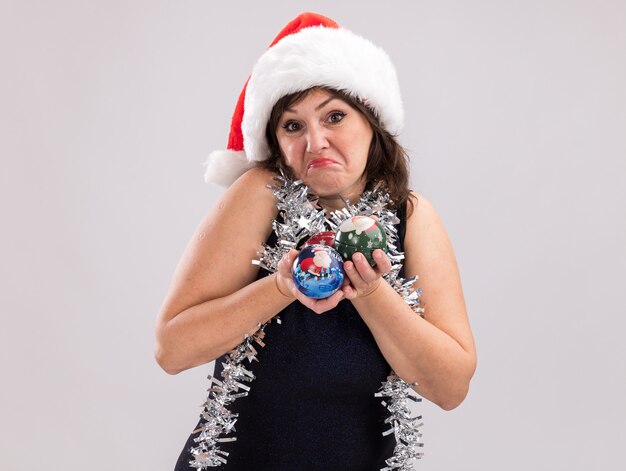 Femme d'âge moyen confuse portant un bonnet de noel et une guirlande de guirlandes autour du cou tenant des boules de noël regardant la caméra isolée sur fond blanc avec espace de copie