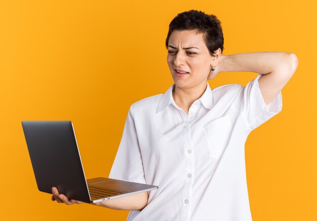 Femme d'âge moyen confuse tenant et regardant un ordinateur portable en gardant la main derrière la tête