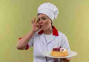 Photo gratuite femme d'âge moyen cuisinier en uniforme de chef tenant un gâteau sur une assiette et montrant un geste délicieux sur un mur vert isolé