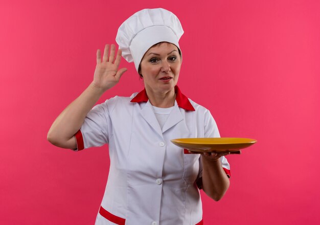 Femme d'âge moyen cuisinier en uniforme de chef tenant la plaque donner bonjour sur un mur rose isolé avec copie espace