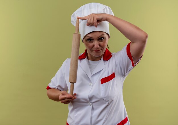 Photo gratuite femme d'âge moyen cuisinier en uniforme de chef tenant le rouleau à pâtisserie avec ses doigts sur mur vert isolé