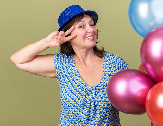 Femme d'âge moyen drôle et joyeuse en chapeau de fête avec un tas de ballons colorés montrant le signe v