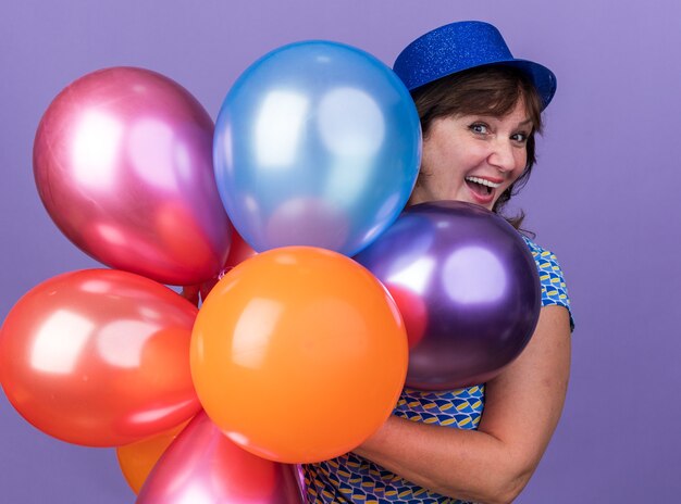 Femme d'âge moyen heureuse et excitée en chapeau de fête avec bouquet de ballons colorés souriant célébrant la fête d'anniversaire debout sur un mur violet
