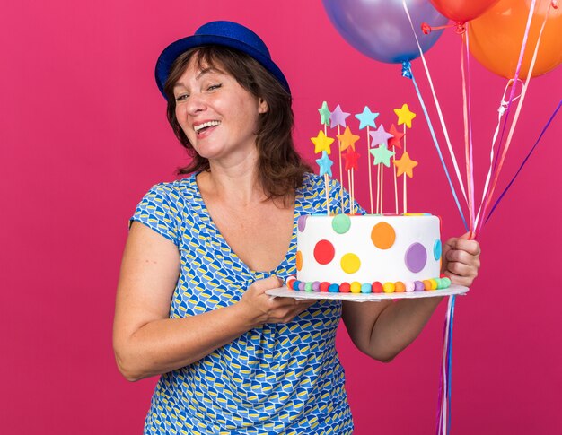 Femme d'âge moyen heureuse et gaie en chapeau de fête avec des ballons colorés tenant un gâteau d'anniversaire souriant largement