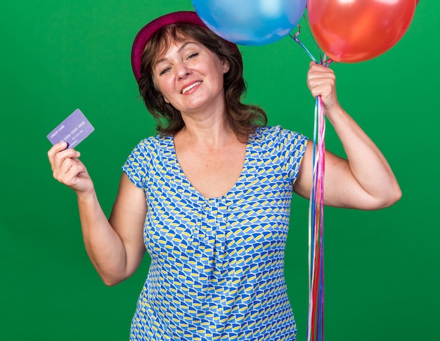 Femme d'âge moyen heureuse et positive en chapeau de fête tenant des ballons colorés et une carte de crédit souriant joyeusement célébrant la fête d'anniversaire debout sur un mur vert