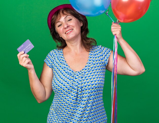 Femme d'âge moyen heureuse et positive en chapeau de fête tenant des ballons colorés et une carte de crédit souriant joyeusement célébrant la fête d'anniversaire debout sur un mur vert