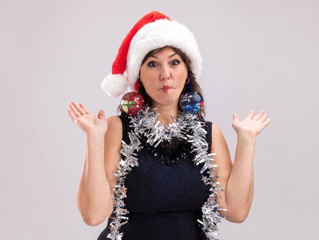 Femme d'âge moyen idiote portant un bonnet de Noel et une guirlande de guirlandes autour du cou regardant la caméra montrant des mains vides pinçant les lèvres avec des boules de Noël suspendues à ses oreilles isolées sur fond blanc