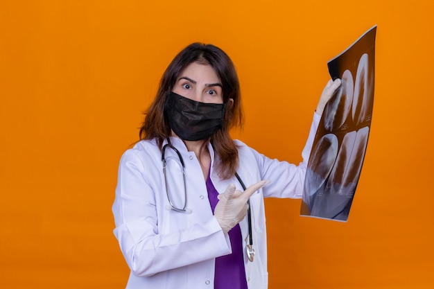 Femme d'âge moyen médecin portant une blouse blanche en masque facial de protection noir et avec stéthoscope holding x-ray des poumons à la surprise en pointant avec l'index à x-ray debout sur isola