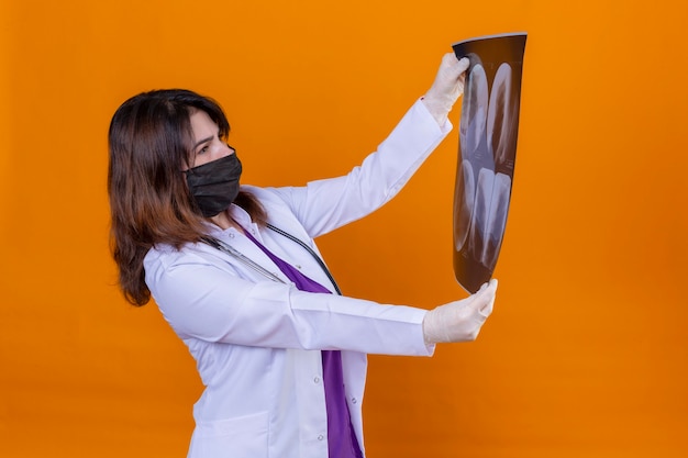 Photo gratuite femme d'âge moyen médecin portant une blouse blanche en masque facial de protection noir et avec stéthoscope tenant des rayons x des poumons à la recherche avec intérêt debout sur fond orange isolé