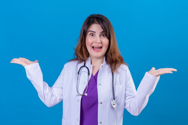 Femme d'âge moyen médecin portant un blouse blanche et avec stéthoscope heureux et sorti debout avec des paumes sur fond bleu