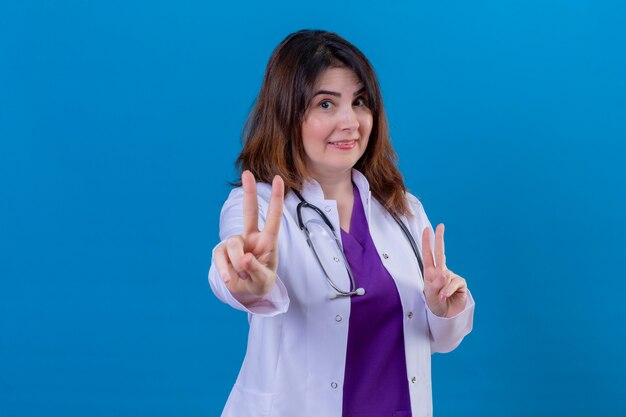 Femme d'âge moyen médecin portant un manteau blanc et avec stéthoscope regardant la caméra en souriant joyeusement faisant signe de la victoire avec les deux mains debout sur fond bleu