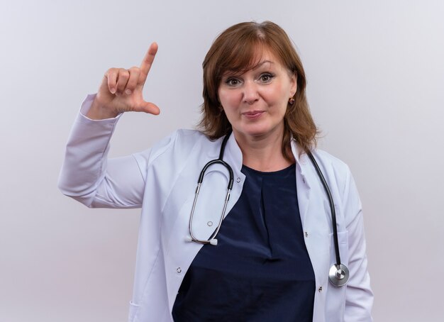 Femme d'âge moyen médecin portant une robe médicale et un stéthoscope faisant un geste de mesure à la recherche sur un mur blanc isolé