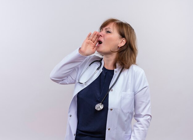 Femme d'âge moyen médecin portant une robe médicale et un stéthoscope mettant la main près de la bouche sur un mur blanc isolé avec espace de copie