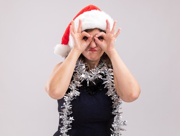 Femme d'âge moyen portant un bonnet de noel et une guirlande de guirlandes autour du cou regardant la caméra faisant un geste de regard en utilisant les mains comme des jumelles isolées sur fond blanc