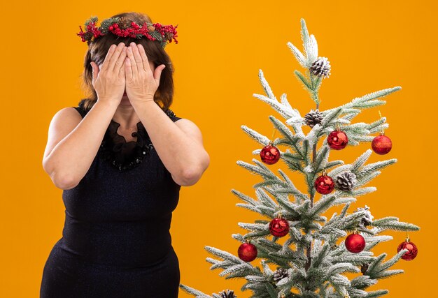Femme d'âge moyen portant couronne de tête de Noël et guirlande de guirlandes autour du cou debout près de l'arbre de Noël décoré couvrant le visage avec les mains isolés sur le mur orange
