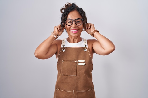 Photo gratuite femme d'âge moyen portant un tablier sur fond blanc souriant tirant les oreilles avec les doigts, geste drôle. problème d'audition