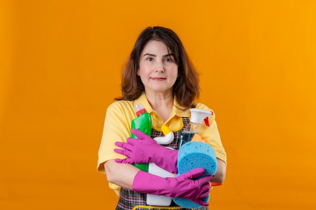 Photo gratuite femme d'âge moyen portant un tablier et des gants en caoutchouc tenant des outils de nettoyage avec une expression sérieuse confiante sur le visage debout sur un mur orange