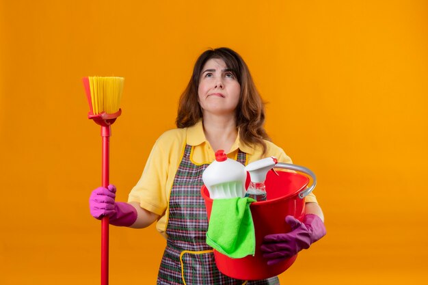 Femme d'âge moyen portant un tablier et des gants en caoutchouc tenant un seau avec des outils de nettoyage et une vadrouille à la recherche avec une expression pensive sur le visage pensant avoir des doutes debout sur un mur orange