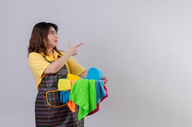 Femme d'âge moyen portant un tablier tenant un seau avec des outils de nettoyage debout sur le côté, pointant vers quelque chose avec le doigt à la surprise sur le mur blanc