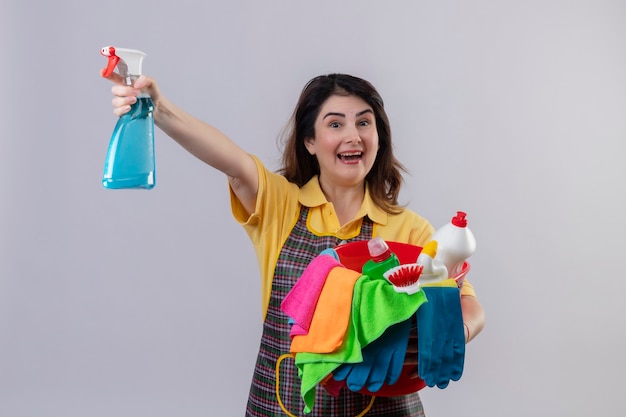 Femme d'âge moyen portant un tablier tenant un seau avec des outils de nettoyage et un spray de nettoyage souriant joyeusement sorti et heureux debout sur un mur blanc