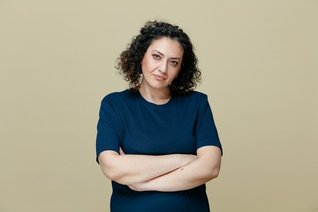 Femme d'âge moyen sérieuse portant un t-shirt regardant la caméra tout en gardant les bras croisés isolé sur fond vert olive