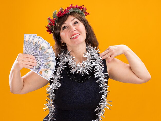 Femme d'âge moyen souriante portant couronne de tête de Noël et guirlande de guirlandes autour du cou tenant de l'argent touchant l'épaule en levant isolé sur fond orange