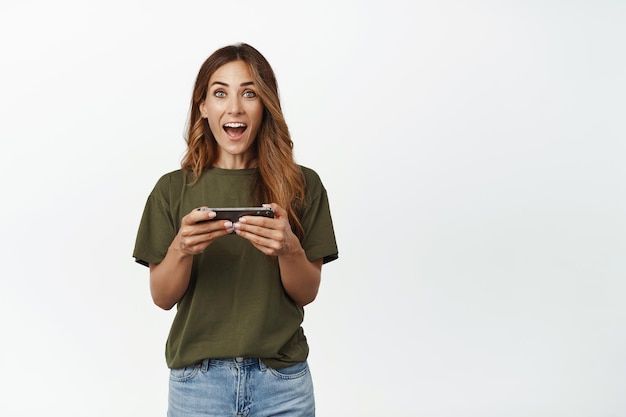 Une femme d'âge moyen surprise et excitée réagit à un jeu vidéo super cool, tenant un téléphone portable horizontalement et souriant étonné, debout sur fond blanc.