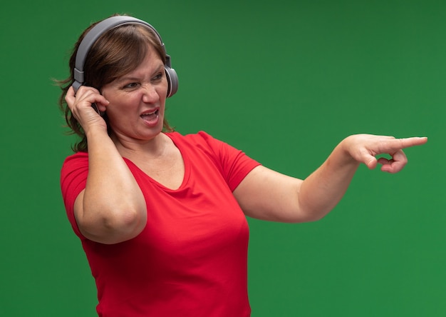 Femme d'âge moyen en t-shirt rouge avec des écouteurs à côté d'être mécontent de pointer avec l'index sur quelque chose debout sur le mur vert