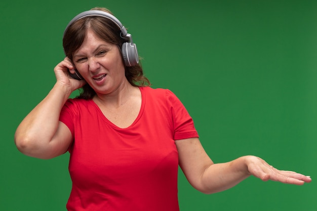 Femme d'âge moyen en t-shirt rouge avec des écouteurs avec une expression dégoûtée en levant le bras debout sur un mur vert