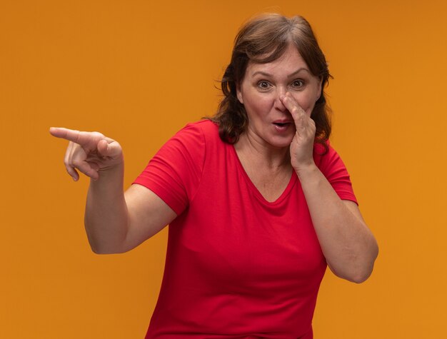 Femme d'âge moyen en t-shirt rouge souriant disant un secret avec la main près de la bouche pointant avec l'index à quelque chose debout sur le mur orange