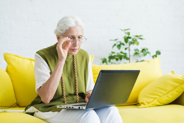 Une femme âgée assise sur un canapé jaune regardant un ordinateur portable