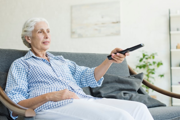 Une femme âgée assise sur un canapé en train de changer de chaîne avec une télécommande