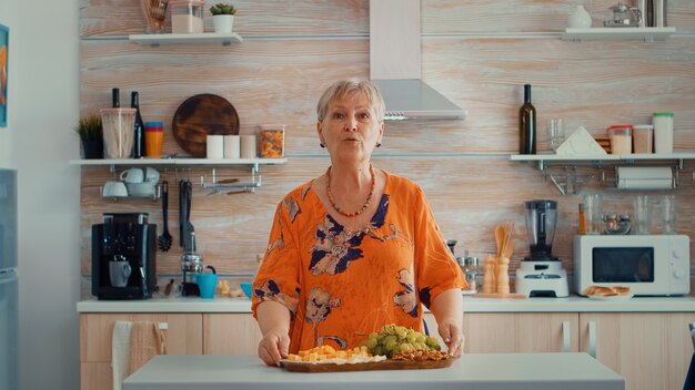 Femme âgée ayant un appel vidéo dans la cuisine pendant que sa famille prépare le dîner en arrière-plan. Pov conférence internet moderne en ligne, chat, communication, appel de conversation par chat via webc