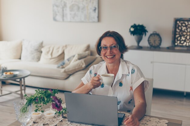 Femme âgée, boire du café et souriant dans la maison en chemise blanche pendant la journée