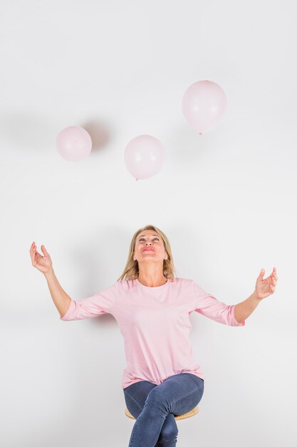 Femme âgée en chemisier rose avec les mains levées sur le tabouret et ballons volants