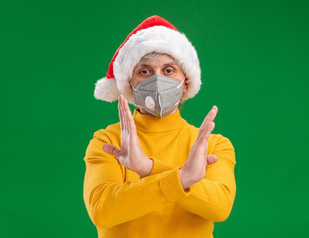 Femme âgée confiante avec Bonnet de Noel portant un masque médical ne faisant aucun signe avec les mains croisées isolé sur fond vert avec espace copie