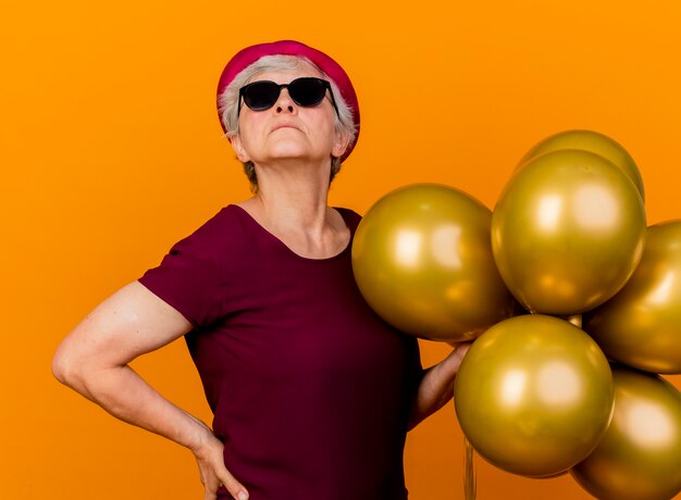 Femme âgée confiante dans des lunettes de soleil portant chapeau de fête se dresse avec des ballons d'hélium isolés sur un mur orange avec espace de copie