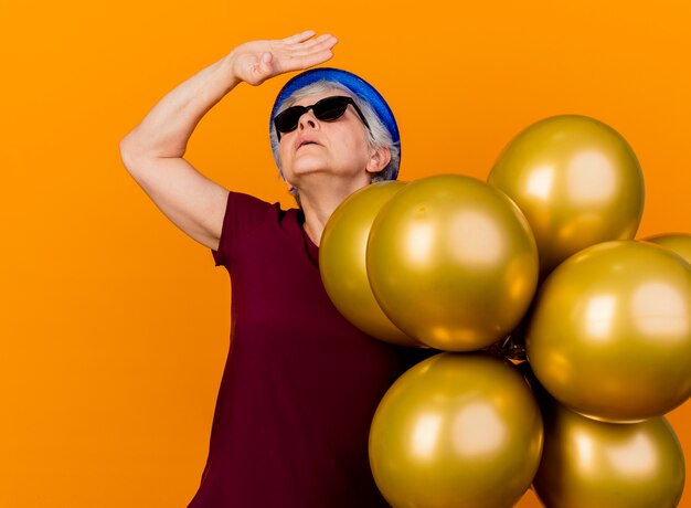 Femme âgée confiante dans des lunettes de soleil portant chapeau de fête se dresse avec des ballons d'hélium en levant la main et en levant isolé sur un mur orange avec copie espace