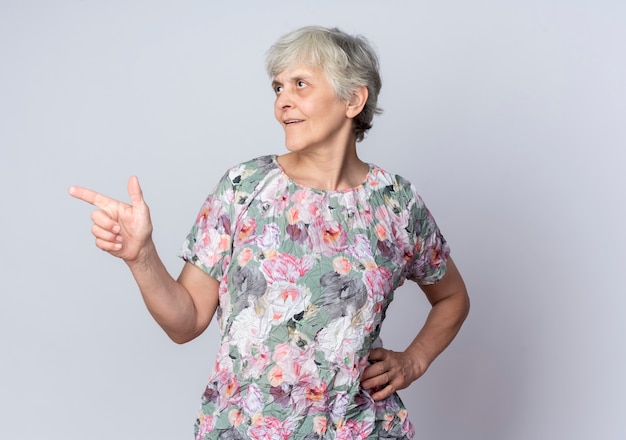 Femme âgée confiante met la main sur la taille à la recherche et pointant sur le côté isolé sur un mur blanc
