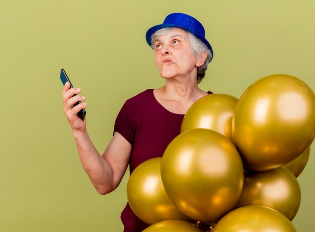 Femme âgée confuse portant chapeau de fête se dresse avec des ballons d'hélium tenant le téléphone en levant isolé sur mur vert olive avec espace copie