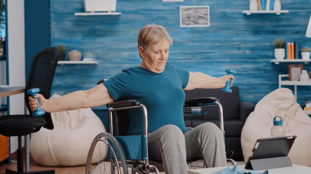 Femme âgée étirant les muscles des bras avec des haltères, assise en fauteuil roulant et regardant la vidéo de la leçon d'entraînement sur tablette. personne âgée ayant un handicap physique utilisant des poids pour faire de l'exercice.