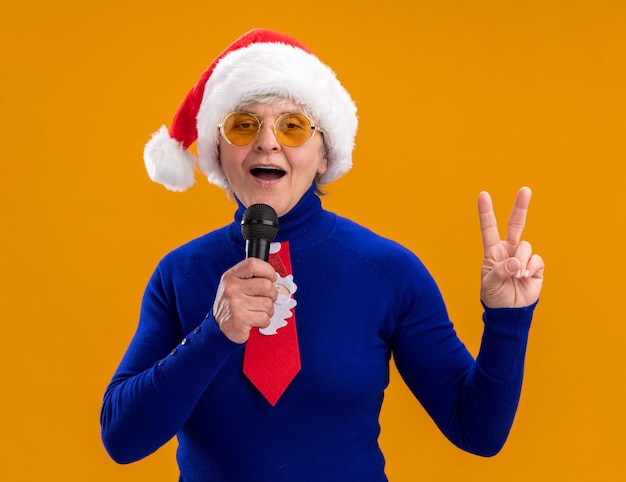 Une Femme âgée Heureuse Dans Des Lunettes De Soleil Avec Un Bonnet De Noel Et Une Cravate De Noel Tient Un Micro Faisant Semblant De Chanter Et Des Gestes Signe De Victoire Isolé Sur Un Mur Orange Avec Espace De Copie