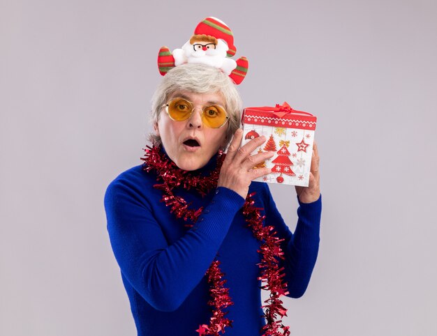 Une femme âgée impressionnée dans des lunettes de soleil avec un bandeau de père Noël et une guirlande autour du cou tient une boîte-cadeau de Noël près de l'oreille isolée sur un mur blanc avec un espace de copie