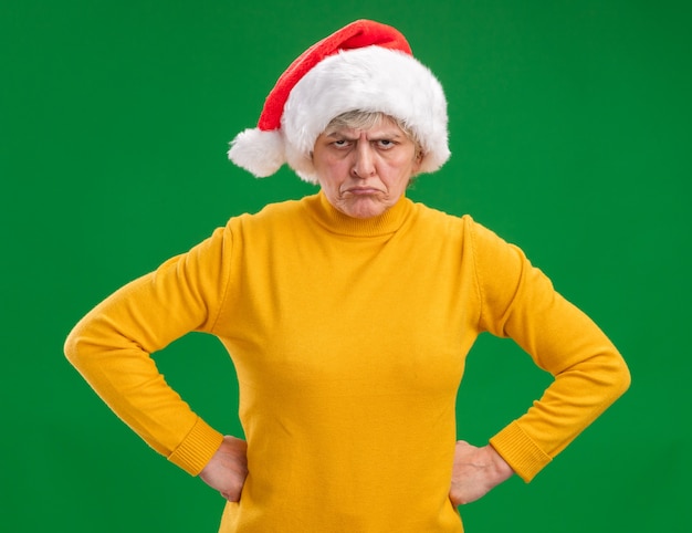 Femme âgée insatisfaite avec Bonnet de Noel met les mains sur la taille et regarde la caméra isolée sur fond violet avec espace copie