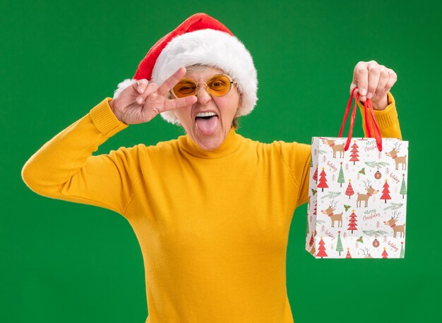 Une femme âgée joyeuse dans des lunettes de soleil avec un bonnet de Noel sort la langue et tient un sac-cadeau en papier gesticulant le signe de la victoire isolé sur un mur vert avec espace pour copie