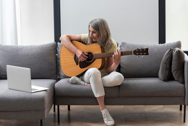 Femme âgée à la maison sur le canapé à l'aide d'un ordinateur portable pour des cours de guitare