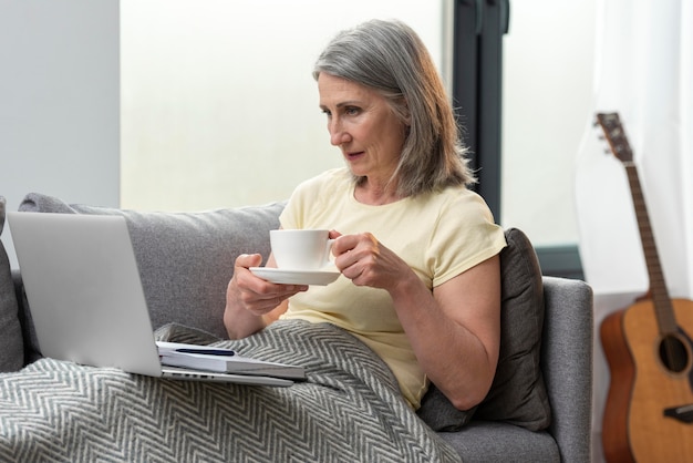 Femme âgée à la maison sur le canapé à l'aide d'un ordinateur portable et prenant un café