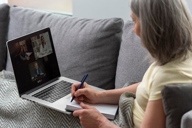 Femme âgée à la maison sur le canapé à l'aide d'un ordinateur portable et prenant des notes