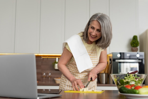 Femme âgée à la maison dans la cuisine prenant des cours de cuisine sur ordinateur portable